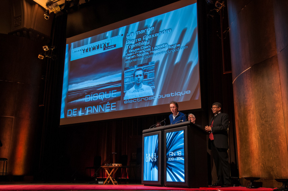 Jean-François Denis is receiving (on behalf of Pierre Alexandre Tremblay visible on the big screen) the Prix Opus 2013-14 “Album of the year — actuelle and electroacoustic musics” at the 18th Gala des Prix Opus at Salle Bourgie in Montréal. The hosts of the event are Pierre Vachon and Stanley Péan [Photo: Anis Hammoud — CQM, Montréal (Québec), February 1, 2015]